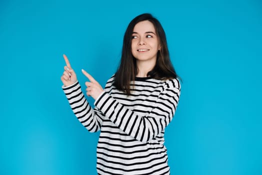 Adorable Girl in Striped Pullover Pointing and Gazing at Empty Space - Charming Young Female Model with Expressive Gestures - Isolated on Blue Background - Perfect for Conceptual, Advertising.