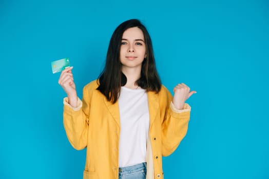 Happy Woman Pointing Thumb at Empty Space with Credit Card - Positive and Attractive Female Expressing Satisfaction and Financial Confidence, Isolated on Blue Background - Ideal for Advertising