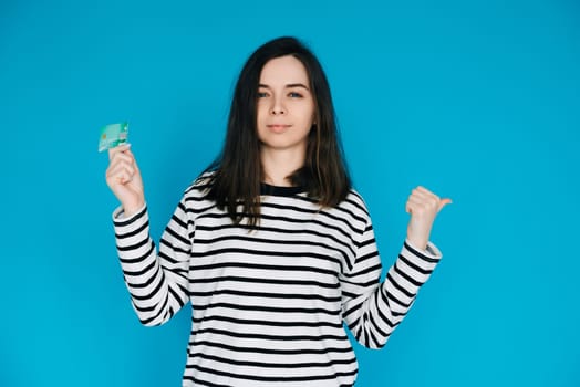 Woman Holding Credit Card, Pointing with Thumb in Empty Space - Female Expressing Confidence and Financial Freedom - Blue Background - Perfect for Banking, E-commerce, and Financial Services Concepts.