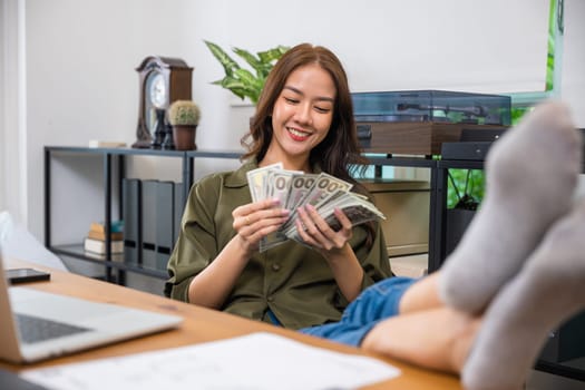 Young woman sitting relaxing reclining with feet up on computer desk counting bunch dollar bills on hands, smiling woman holding money banknotes with laptop, Achievement career, Loan, trading, lottery