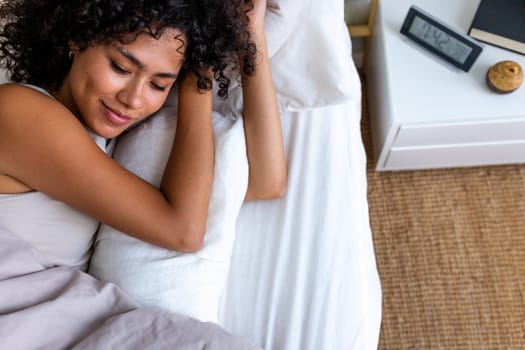 Serene and beautiful young woman with curly hair relaxing, lying down in bed in the morning. Copy space. Lifestyle concept