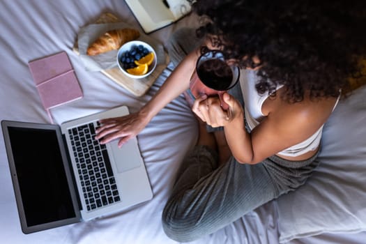 High angle view close-up of young unrecognisable woman sitting on bed typing on laptop while having morning coffee and eating breakfast. Lifestyle concept.