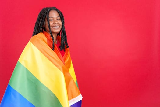 Happy african woman wrapped with a lgbt flag in studio with red background