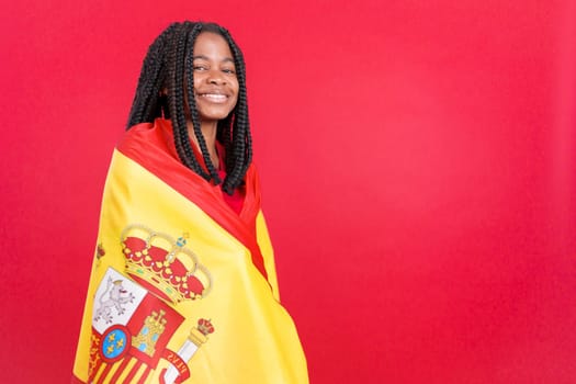 Happy african woman wrapping with a spanish national flag in studio with red background