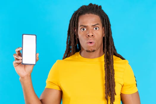 Surprised man with dreadlocks holding a mobile phone with blank screen in studio with blue background