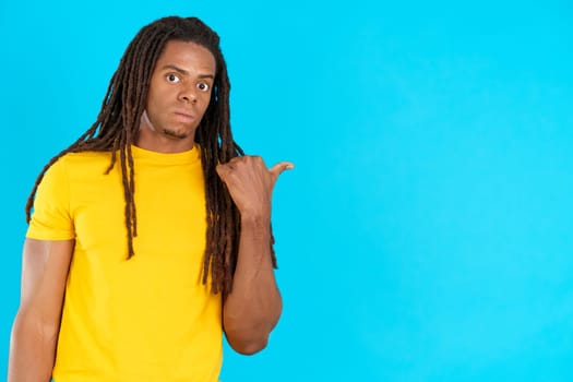 Surprised latin man with dreadlocks pointing aside in studio with blue background