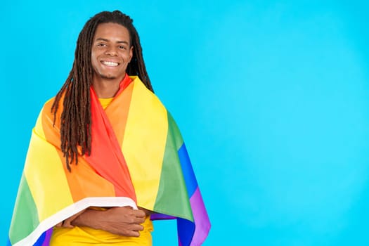 Happy latin man with dreadlocks wrapped with a lgbt flag in studio with blue background