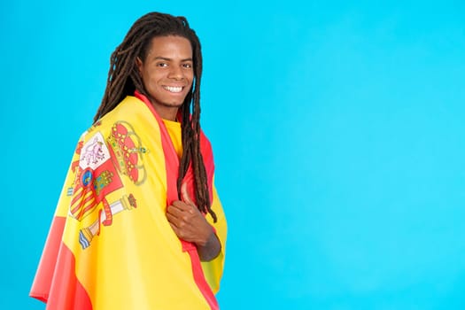 Happy latin man with dreadlocks wrapping with a spanish national flag in studio with blue background