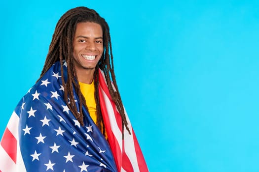 Happy latin man with dreadlocks wrapping with a north america national flag in studio with blue background