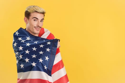 Cheerful gay man wrapping with a north america national flag in studio with yellow background