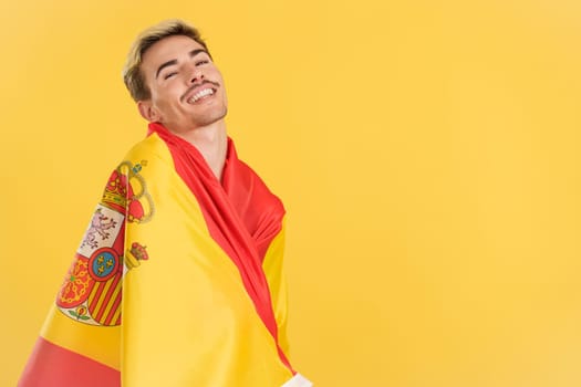 Happy gay man wrapping with a spanish national flag in studio with yellow background