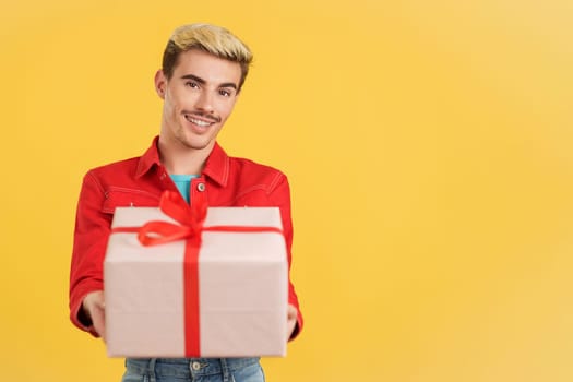 Happy gay man looking at the camera giving a present in studio with yellow background