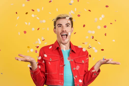 Happy gay man surrounded by confetti flying in the air in studio with yellow background