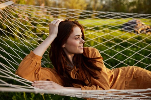 a happy woman is resting in a mesh hammock with her head resting on her hand, smiling happily looking away, enjoying a warm day in the rays of the setting sun, lying in an orange dress. High quality photo