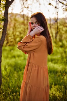 a sweet, thoughtful woman stands in nature near a tree in a long orange dress, illuminated from the back by the sunset rays of the sun and holding her hand near her face looks towards the camera. High quality photo