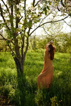 a beautiful, joyful woman stands in a long orange dress near a tree blooming with white flowers during sunset, illuminated from the back and holding her hands near her head looking away. High quality photo