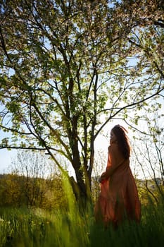 vertical photo of a silhouette of a woman standing in a long orange dress near a flowering tree. High quality photo