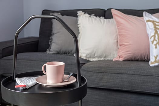 Coffee table with cup of tea, near dark gray sofa with pillows in a living room.