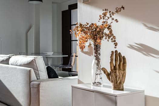 Decorative hand sculpture and vase with flowers on small white cupboard in sunny interior of modern apartment.