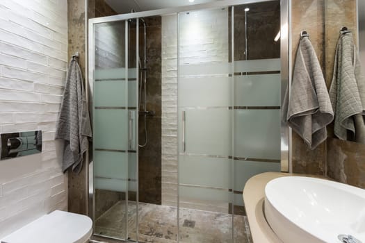 Bathroom in modern style with textured tiles on the floor and walls. Two showers with glass door, white toilet and towels