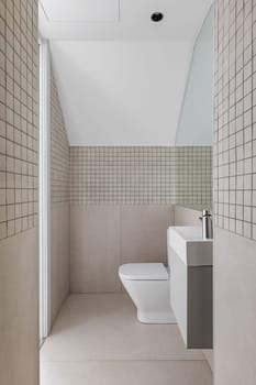 Interior of narrow bathroom with small sink and toilet. View from the shower zone of modern bathroom with beige tiles.