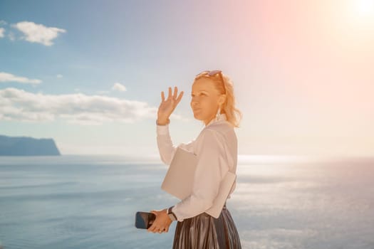 Business woman on nature in white shirt and black skirt. She works with an iPad in the open air with a beautiful view of the sea. The concept of remote work