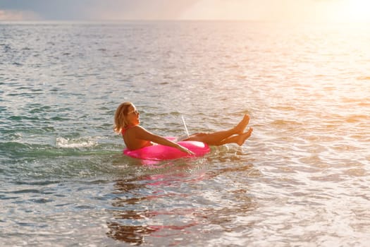 Woman works on laptop in sea. Freelancer, blond woman in sunglases floating on an inflatable big pink donut with a laptop in the sea at sunset. People summer vacation rest lifestyle concept