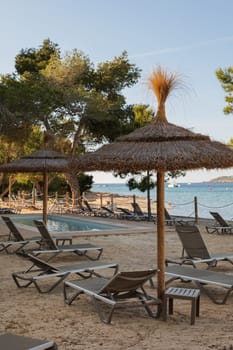 Sunbeds and thatched sunshades on the beach of a hotel with swimming pool and view to the sea on Ibiza island. Beautiful relax zone with the evening light