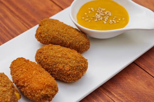 Fried croquettes with yellow sauce on white plate. Side view on wooden background. Traditional spanish snack.
