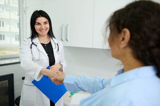 Confident Caucasian woman, female doctor, GP,physician, gynecologist holding clipboard with medial insurance contract, smiling broadly and shaking hand while welcoming a new patient in her office