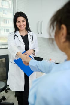 Charming pleasant female doctor, GP,physician, gynecologist holding clipboard with medial insurance contract, smiling broadly and shaking hand while welcoming a gravid pregnant patient in her office