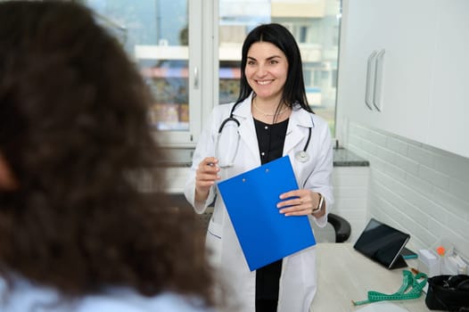 Confident portrait of a Caucasian charming pretty woman, female doctor, GP, physician, gynecologist in white medical gown, smiling broadly, welcoming her patient at appointment in new medical clinic