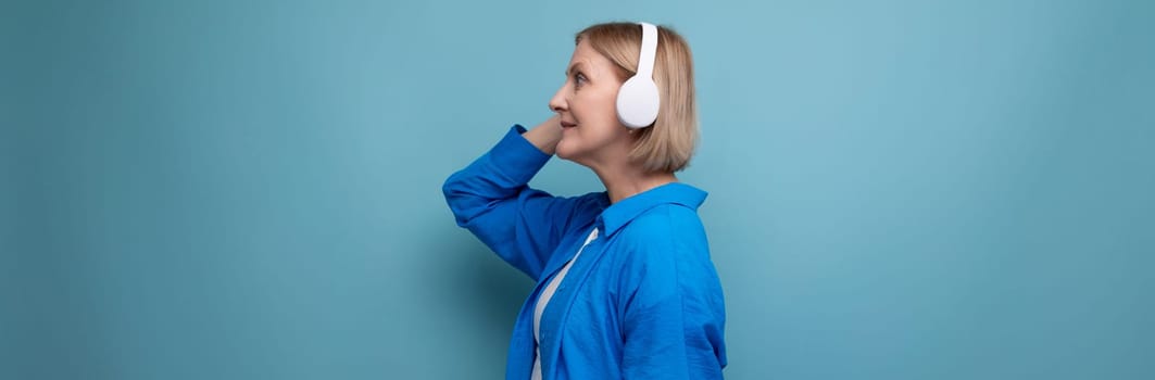 middle age business concept. mature woman listening to audiobook with headphones on blue background copy space.