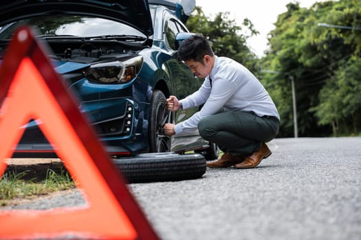 Asian businessman car broken has problems with wheel of his car during go to work in morning he changing replace rubber tire on wheels, business man have warning sign on road, problem transportation