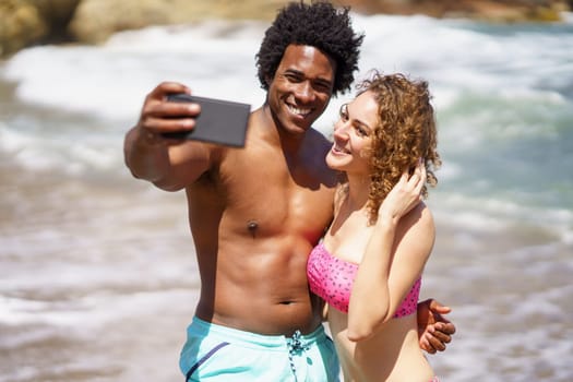 Happy young multiethnic couple in swimwear hugging while taking selfie on mobile phone for social media during honeymoon