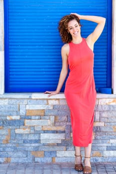 Full body of young female in red sundress and flat sandals, standing on street and looking at camera while touching hair and leaning on hand on wall against blue shutters