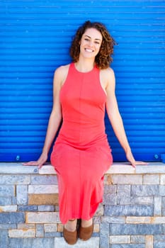 Full body of relaxing young female in red sundress sitting with hands on stonewall and smiling while looking at camera against blue shutters in daylight