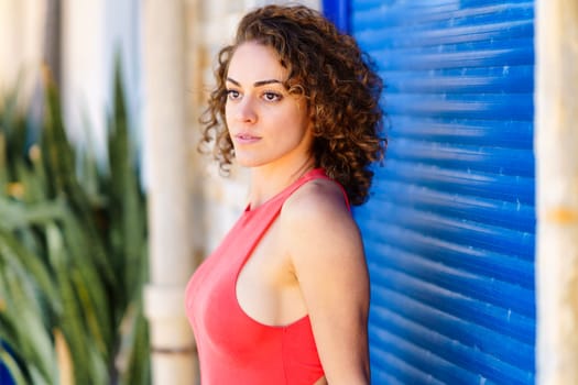 Side view of beautiful young female in red sleeveless sundress standing in front of building with blue shutters and looking away against blurred tropical plants