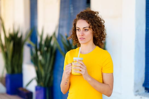 Thoughtful young curly haired female in yellow t shirt standing on street with glass jar of orange juice and looking away dreamily