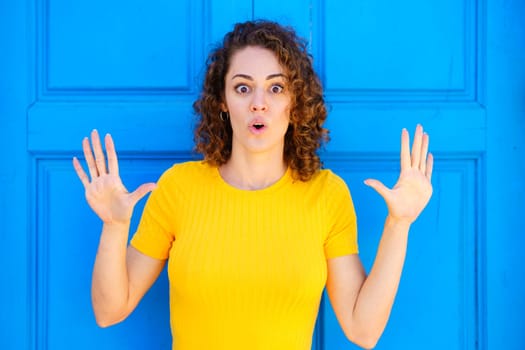 Shocked young female in yellow t shirt, with curly hair and opened mouth looking at camera with raised hands while standing against blue wall on street