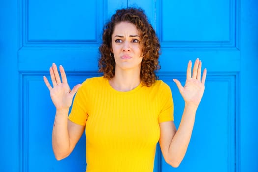 Scared young female in yellow t shirt with curly brown hair looking away with raised hands against blue background on city street