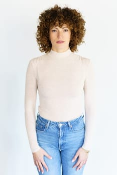 Adult female in beige turtleneck and jeans with brown curly hair looking at camera while standing still with arms hanging straight on sides