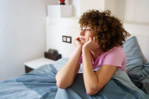 Hispanic curly woman leaning on arms and looking away thoughtfully while sitting on bed after getting up