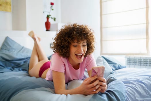 Full body of amazed young woman with curly short hair lying on comfortable bed while watching video on mobile phone in bedroom at home