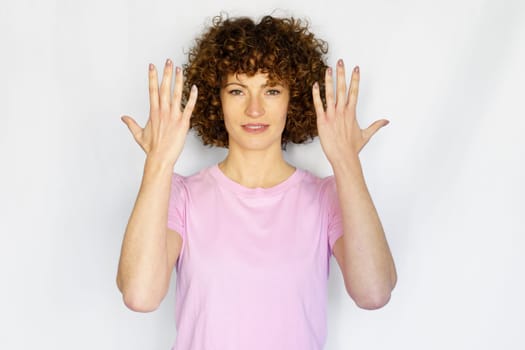 Positive curly haired woman showing number ten on fingers while raising arms and looking at camera against white background