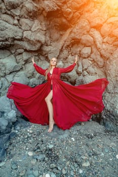 Red dress rocks woman. A blonde with flowing hair in a long flowing red dress stands near a rock of volcanic origin. Travel concept, photo session at sea.