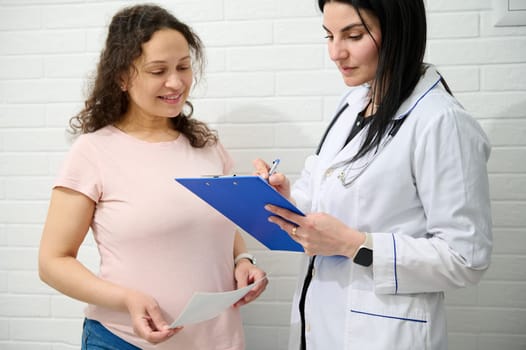 Positive pregnant woman at obstetrician appointment. Female doctor gynecologist with clipboard consulting a gravid expectant mother, examining patient and prescribing medicines. Pregnancy. 9 month