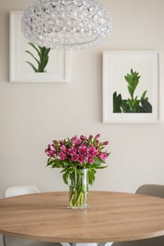 Vase of flowers stands on a wooden table on the background of wall with posters of green plants with beige wallpaper with crystal chandelier. Concept of concise and stylish interior design.