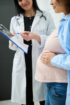 Cropped view of a pregnant woman holding her hand on her big belly, smiling while listening to her doctor explaining the ultrasound scan of her baby, checking her health and prescribing medicines