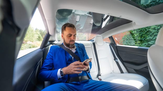 A caucasian man in a blue suit uses a smartphone in the back seat of a car. Business class passenger